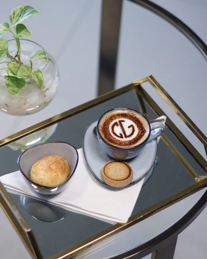 Cappuccino com o monograma da clínica na espuma, pão de queijo e biscoito em bandeja dourada.