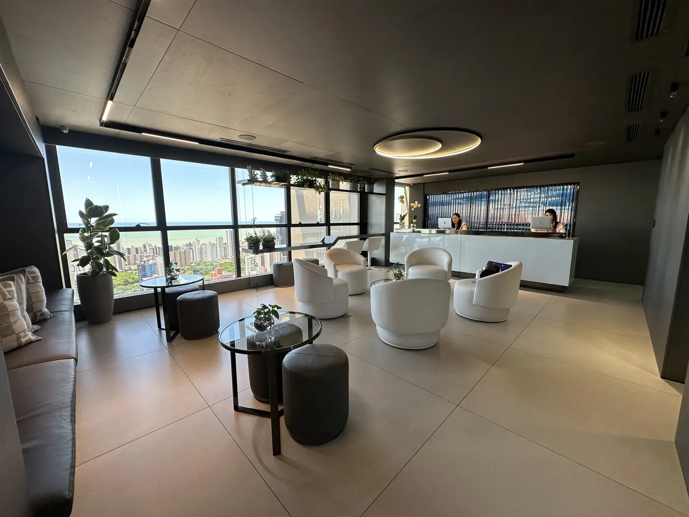 Recepção e lounge da clínica, poltronas brancas, balcão de atendimento e janelas com vista para a cidade e o mar.